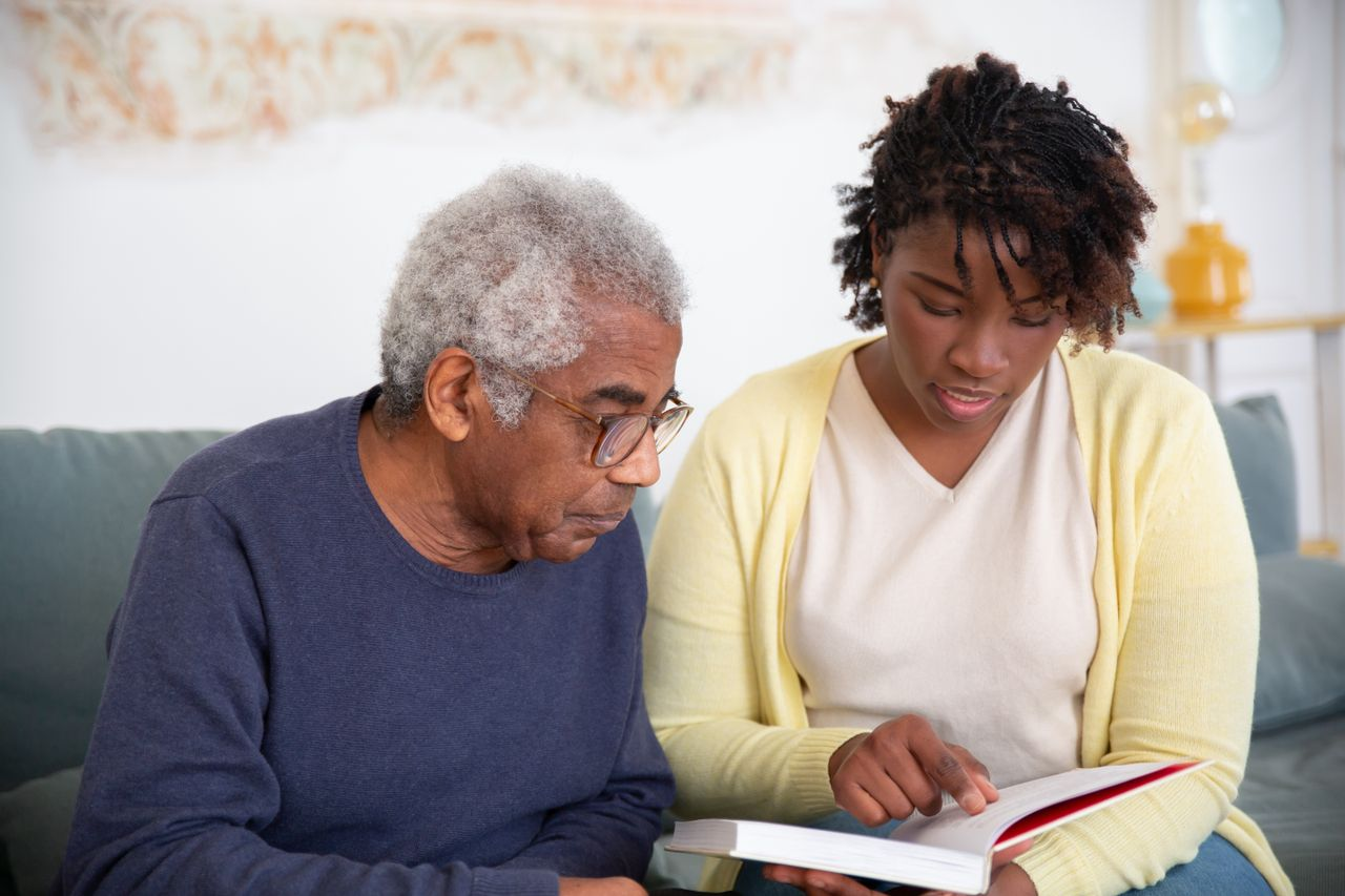 A caregiver helps a senior man with reading, providing support at home.